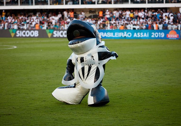 Mascote do Santos Futebol Clube em campo durante jogo oficial, interagindo com a torcida